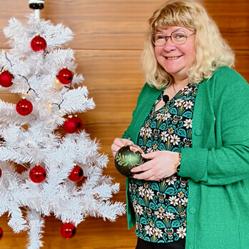 Eine Frau steht mit einer grünen Weihnachtskugel in der Hand vor einem weißen geschmückten plastik Weihnachtsbaum